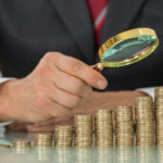 Close-up Of Businessman Hand Holding Magnifier On Coin At Desk